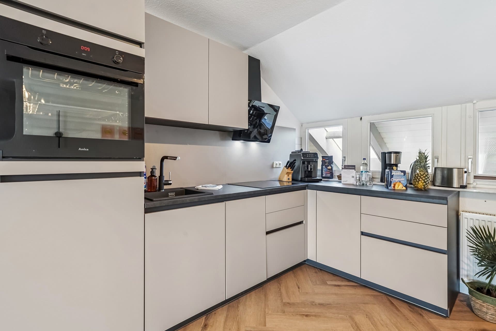 Fitted kitchen under a sloped ceiling with herringbone parquet; the dormer windows stay detailed despite the backlight